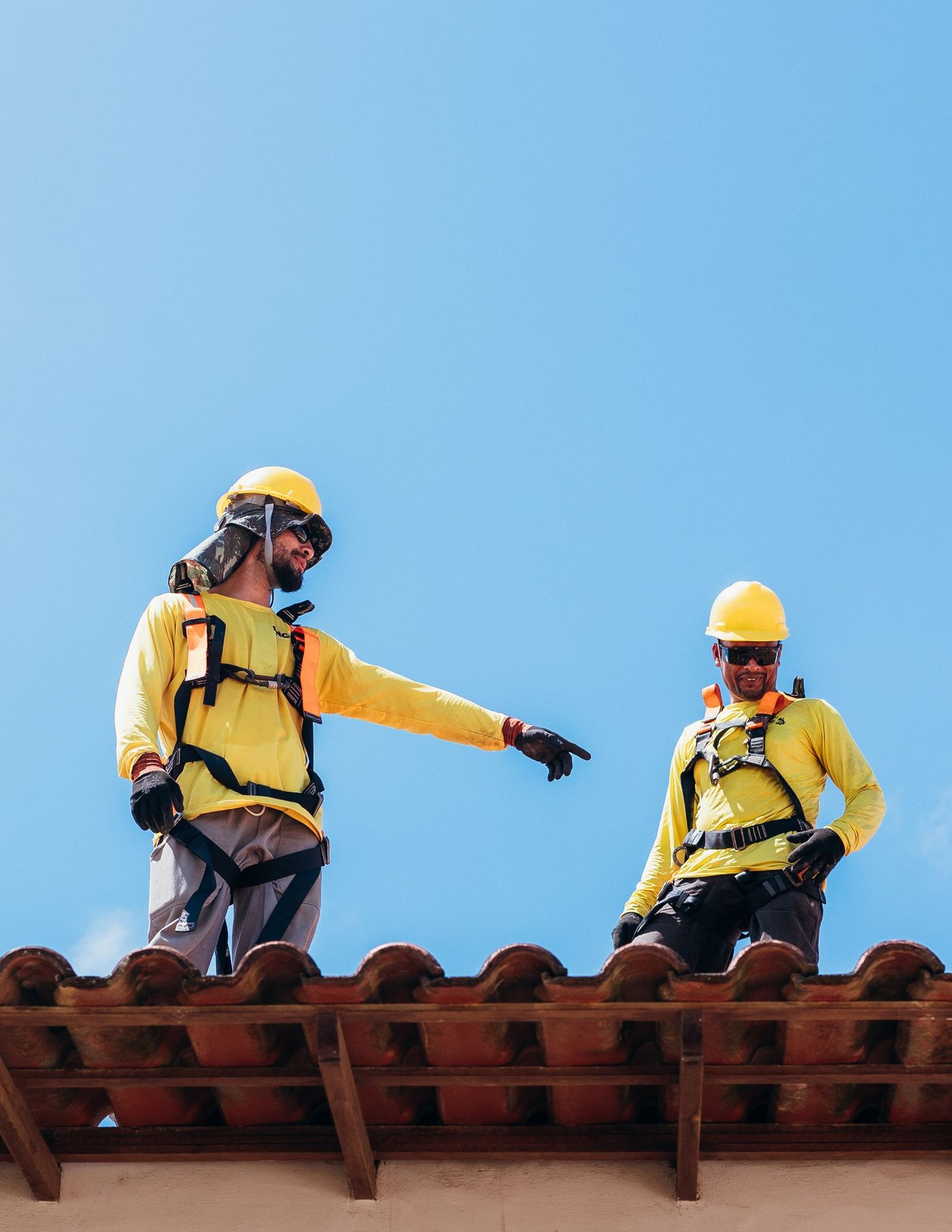 Two workers in harnesses and hard hats standing on a tiled roof, one pointing across the roof