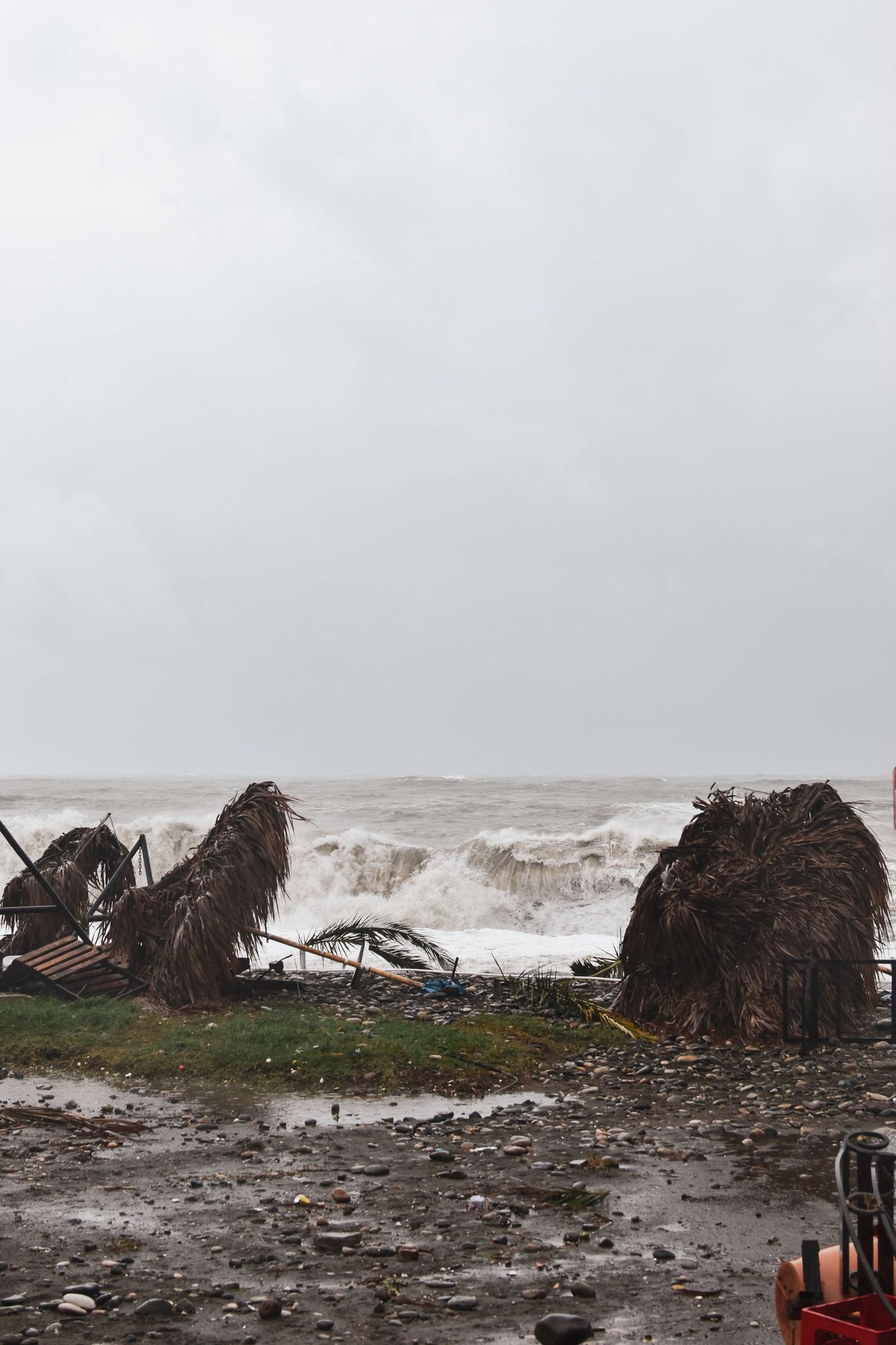Beachfront after a storm: torn palm-thatch shelters, debris on the ground and heavy surf under a grey sky